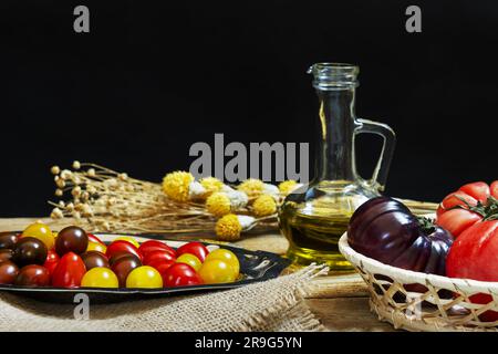 Einfaches Stillleben mit reifen Tomaten verschiedener Sorten und kleinen Kirschtomaten verschiedener Farben zusammen mit Olivenöl und getrockneten Blumen Stockfoto
