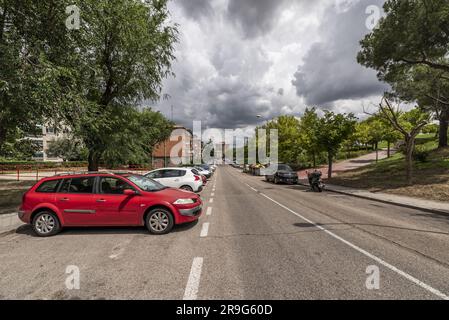 Eine Straße mit einem städtischen Park auf der einen Seite und Autos, die an einem Tag mit einem Brausturm am Himmel geparkt werden Stockfoto