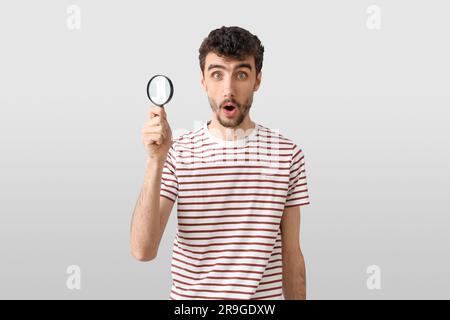 Schockierter junger Mann mit Lupe auf hellem Hintergrund Stockfoto