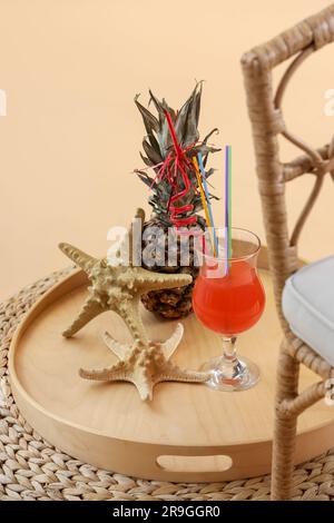 Holztablett mit Cocktail, Ananas und Seesternen auf Korbottomane vor hellorangefarbenem Hintergrund. Reisekonzept Stockfoto