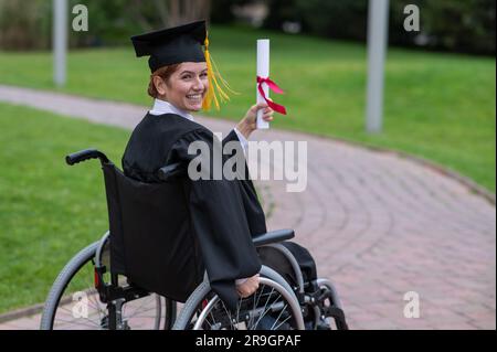 Glückliche weiße Frau im Rollstuhl dreht sich um und hält ihr Diplom draußen. Stockfoto