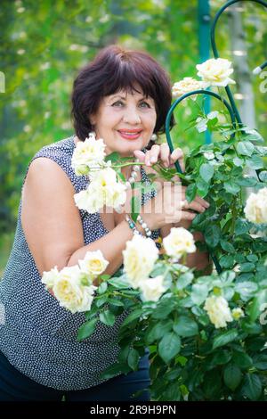 66-jährige Frau im Garten. Eine reife Frau genießt die Blumen einer Rose, die mit ihren eigenen Händen in ihrem Garten gewachsen ist. Stockfoto