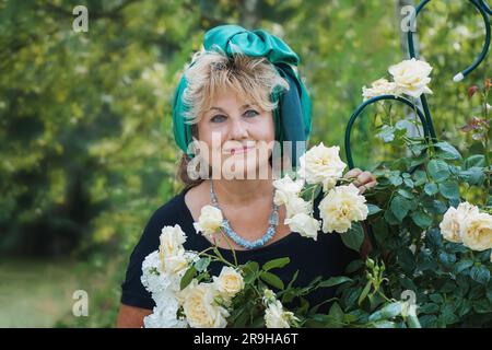 59-jährige Frau im Garten. Eine reife Frau genießt die Blumen einer Rose, die mit ihren eigenen Händen in ihrem Garten gewachsen ist. Stockfoto