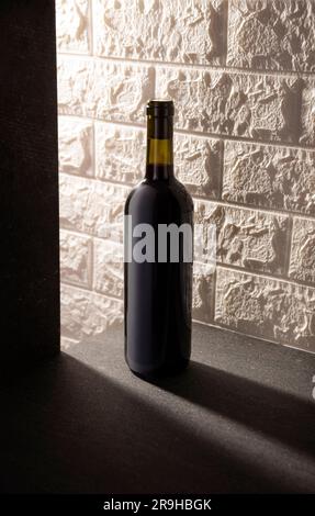 Unetikettierte Rotweinflasche auf grauer Steinoberfläche, weißer rustikaler Backsteinhintergrund. Diagonale lange Schatten. Minimalistisches Konzept. Stockfoto