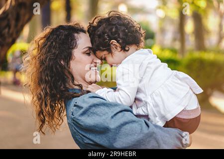 Eine hellbraune, gelockte Mutter hält ihre 3-jährige Tochter, deren Stirn sich berührt und lacht, gefangen im Profil eines öffentlichen Parks. Stockfoto