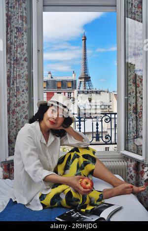 Frau, die auf einem Bett in einem Hotelzimmer sitzt und einen Fensterblick auf den Eifelturm hat, eine reife asiatische Touristin in Paris. Stockfoto