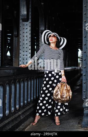 Eine stilvolle, glamouröse reife Frau, umgeben von Eisensäulen unter Pont Alexandre III Paris, mit Streifen und Polka Dots, Schwarz, weiß und Gold Stockfoto