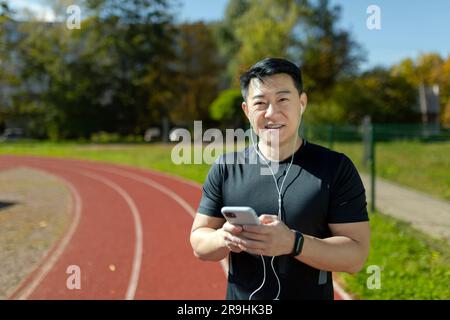 Porträt eines erfolgreichen asiatischen Mannes, der im Freien joggt und sich sportlich betätigen kann. Läufer, der mit dem Telefon in den Händen und Kopfhörern in die Kamera schaut, Musik hört und Online-Podcasts anhört. Stockfoto