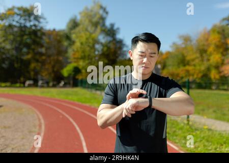 Asiatischer Läufer überprüft Herzfrequenz und Fortschritt auf Fitness Armband Smartwatch, seriöser Mann sieht aufmerksam aus, läuft und trainiert im Stadion. Stockfoto