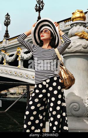 Eine modische Asiatin mittleren Alters in Paris unter Pont Alexandre III, die ein gestreiftes bretonisches T-Shirt und Hosenröcke mit Punktmuster trägt. Stockfoto