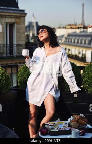 Eine modische, dunkelhaarige asiatische Frau in weißem Hemdkleid auf der Terrasse des Hotelzimmers beim Frühstück mit Blick auf Paris und den Eiffelturm Stockfoto