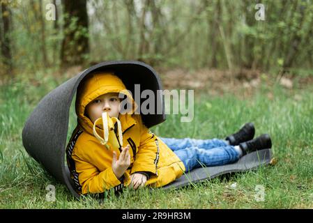 Ein kleiner Junge ruht auf einer Touristenmatte im Wald und isst eine Banane. Stockfoto