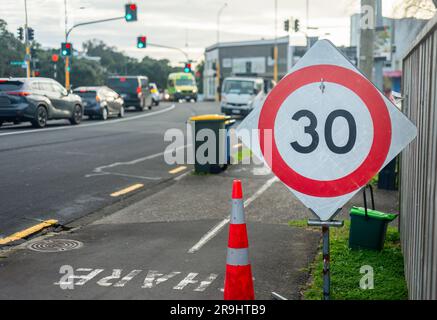 30km Geschwindigkeitsbegrenzungsschild und Mülltonnen am Straßenrand. Autos an der Kreuzung. Auckland. Stockfoto