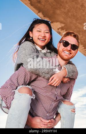 Vertikales Porträt eines gutaussehenden Freundes mit Sonnenbrille, der seiner freudigen Freundin einen Huckepack gibt. Ein junges, multiethnisches, glückliches Paar, das Spaß miteinander hat und bei einem romantischen Date vor die Kamera schaut. Hochwertiges Foto Stockfoto
