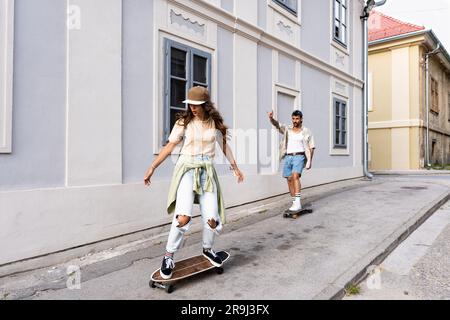 Touristen Paare Skateboarder, die Schlittschuhe fahren. Glückliche junge Reisende, die gemeinsam auf dem Asphalt bei Sonnenuntergang sitzen. Stylischer Mann und Frau in trendigen Outfits Stockfoto