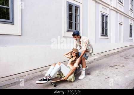 Touristen Paare Skateboarder, die Schlittschuhe fahren. Glückliche junge Reisende, die gemeinsam auf dem Asphalt bei Sonnenuntergang sitzen. Stylischer Mann und Frau in trendigen Outfits Stockfoto