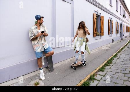 Touristen Paare Skateboarder, die Schlittschuhe fahren. Glückliche junge Reisende, die gemeinsam auf dem Asphalt bei Sonnenuntergang sitzen. Stylischer Mann und Frau in trendigen Outfits Stockfoto