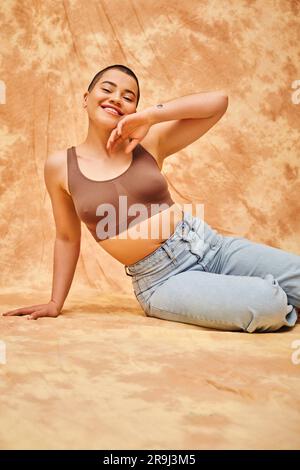 Körperliebe, Jeans, kurvige und tätowierte Frau in legerer Kleidung, die auf einem fleckigen beigen Hintergrund sitzt, Selbstvertrauen, Selbstakzeptanz, Generation z, Körper Stockfoto