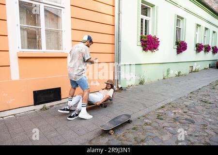 Touristen Paare Skateboarder, die Schlittschuhe fahren. Glückliche junge Reisende, die gemeinsam auf dem Asphalt bei Sonnenuntergang sitzen. Stylischer Mann und Frau in trendigen Outfits Stockfoto