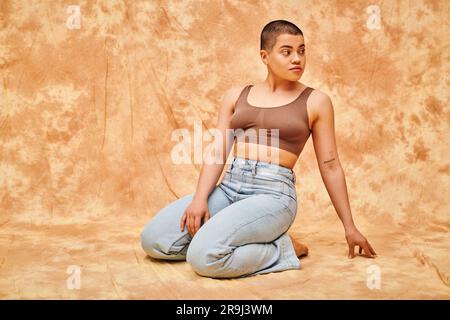 Körperpositivität, Jeansmode, kurvige und tätowierte Frau in Jeans und Crop Top auf einem fleckigen beigen Hintergrund, lässige Kleidung, wegschauen, selbst-- Stockfoto