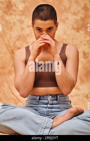 Positive Körperhaltung, kurvige und tätowierte Frau in Jeans und Crop Top sitzend mit gekreuzten Beinen auf meliertem beigefarbenem Hintergrund, schauend auf die Kamera, persönlicher Stall Stockfoto