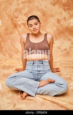 Körperpositivität, Freizeitkleidung, kurvige junge und tätowierte Frau in Jeans und Crop Top sitzend mit gekreuzten Beinen auf meliertem beigefarbenem Hintergrund, persönliche s Stockfoto