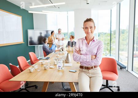 Lächelnde Geschäftsfrau mit gekreuzten Armen, die auf einem Konferenztisch sitzt Stockfoto