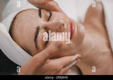 Von oben von einer anonymen Weibchen, die feuchtigkeitsspendende Creme auf das Gesicht aufträgt, während sie zu Hause auf dem Bett in einem hellen Schlafzimmer sitzt Stockfoto