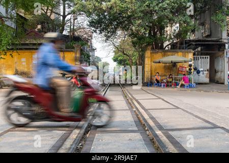 Hanoi, Vietnam-April 2023; Flachblick auf die geschäftige Straße mit Roller in unscharfer Bewegung, der die Bahngleise der berühmten Eisenbahnlinie durchquert Stockfoto