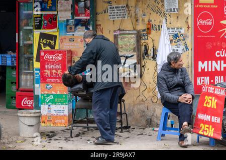 Hanoi, Vietnam-April 2023; Nahaufnahme eines Friseurs, der einen Mann in einem Liegestuhl auf dem Gehweg eines Stree rasiert, während ein anderer Kunde wartet Stockfoto