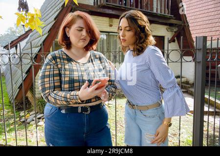 Zwei junge Latina-Frauen argentinischer ethnischer Herkunft, die vor ihrem Haus standen, das Telefon überprüften und über die mobile App ein Taxi rufen. Stockfoto