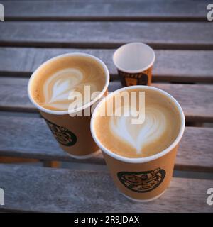 Zwei Papptassen Kaffee mit herzförmigem Latte Art auf verschwommenem Holzhintergrund. Morgens Kaffee zum Mitnehmen für Liebespaare. Stockfoto