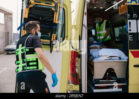 Seitenansicht des Krankenwagenmitarbeiters in Uniform, der mit liegendem Patienten am Auto steht und die Tür während der Arbeit schließt Stockfoto