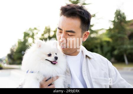 Ein Asiate in lässigen Outfits hält den niedlichen flauschigen Spitz vor dem Hintergrund eines Sommerparks Stockfoto