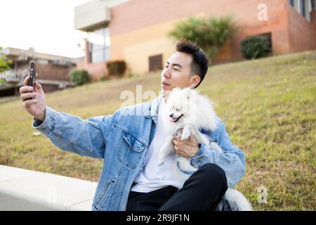 Asiatischer männlicher Hundebesitzer in Freizeitkleidung mit niedlichem Pommern-Spitz und Smartphone, das Selbstporträt auf dem Bürgersteig macht Stockfoto