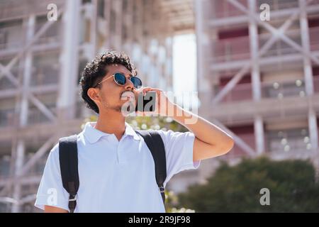 Ein bärtiger Typ in trendiger Sonnenbrille, der bei Sonnenlicht mit seinem Smartphone wegsieht Stockfoto