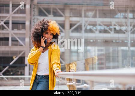 Positive schwarze Frau mit gelben und schwarzen Afro-Haaren in legerer Kleidung. Sie steht an der Stadtstraße neben einem modernen Apartment und telefoniert und schaut nach Stockfoto