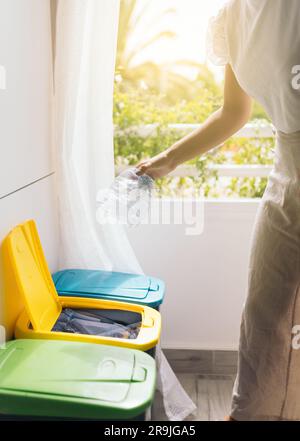 Seitenansicht einer anonymen jungen Erntegutfrau, die eine Plastikflasche in einen gelben Behälter in der Küche in Fensternähe vor verschwommenem Hintergrund wirft Stockfoto