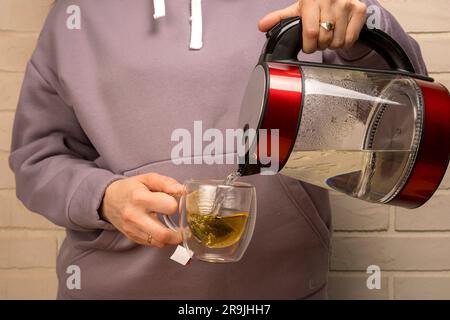 Nahaufnahme einer Frau, die heißes Wasser aus einem Wasserkocher in einen durchsichtigen Glasbecher gießt. Eine Tasse frisch gebrühten Kräutertee. Das Zubereiten von Tee oder Teecere Stockfoto