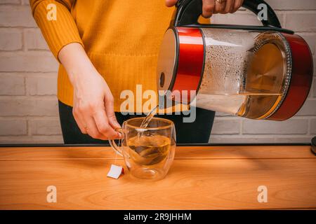Nahaufnahme einer Frau, die heißes Wasser aus einem Wasserkocher in einen durchsichtigen Glasbecher gießt. Eine Tasse frisch gebrühten Kräutertee. Das Zubereiten von Tee oder Teecere Stockfoto