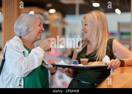 Seitenansicht einer lächelnden alten Mutter und reifer Tochter, die neben der Theke stehen und Kaffee in der Cafeteria trinken Stockfoto