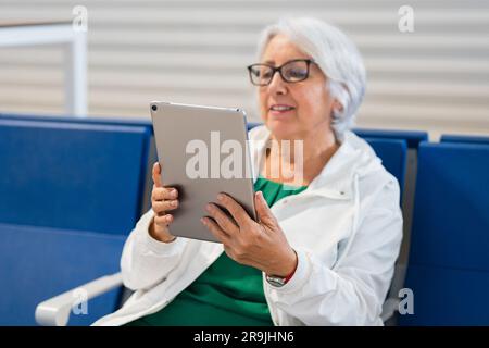 Positive ältere Frau mit kurzen grauen Haaren, die sich einen Film auf einem Tablet ansieht, während sie im Warteraum sitzt Stockfoto