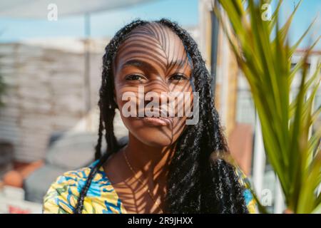 Porträt einer lächelnden jungen afroamerikanischen Frau mit langen Dreadlocks, die in die Kamera schauen, während sie an einem sonnigen Tag neben Palmenblatt steht Stockfoto
