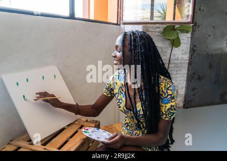 Positive junge afroamerikanische Künstlerin in lässiger Kleidung mit Dreadlocks-Malerei auf Leinwand, während sie im Workshop Bilder kreiert Stockfoto