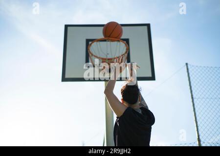 Von unten betrachtet wirft eine anonyme Basketballspielerin den Ball auf dem Sportplatz an sonnigen Tagen in den Korb Stockfoto