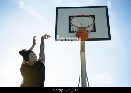 Von unten betrachtet wirft eine anonyme Basketballspielerin den Ball auf dem Sportplatz an sonnigen Tagen in den Korb Stockfoto