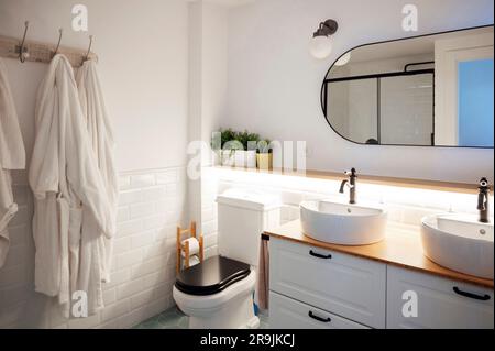 Minimale Innenausstattung des Badezimmers mit Bademänteln auf einem Hängestänge und Wasserspülen auf einem Holzschrank mit Regalen, Spiegel darüber und Toilettenschüssel in der Ecke Stockfoto