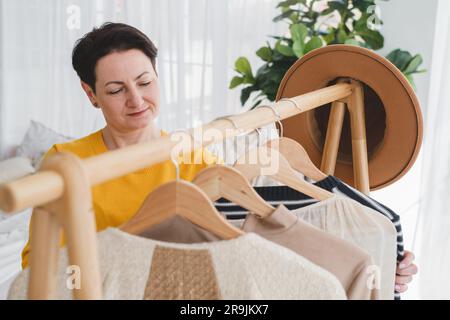 Von oben lächelnde Frau mittleren Alters mit kurzen schwarzen Haaren in gelbem Pullover, die neben einem Holzbügel steht und Kleidung in einem hellen Apartment aussucht Stockfoto