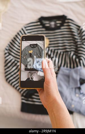 Draufsicht auf eine anonyme Frau, die ein Foto von Kleidung macht, die über ein Smartphone im Schlafzimmer auf das Bett gelegt wurde Stockfoto