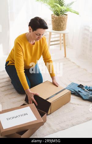 Hochwinkel einer fokussierten Frau mittleren Alters mit kurzen Haaren in Freizeitkleidung, die auf einem Teppich sitzt und Kleidung in Kartonschachteln zur Spende verpackt Stockfoto